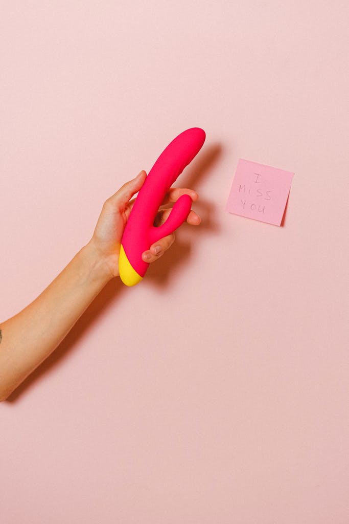 A close-up of a hand holding a pink silicone vibrator next to a post-it note on a pastel backdrop.