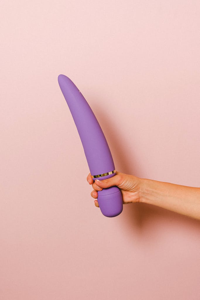 A minimalist shot of a hand holding a purple silicone vibrator against a pink backdrop, highlighting pleasure and sexuality.