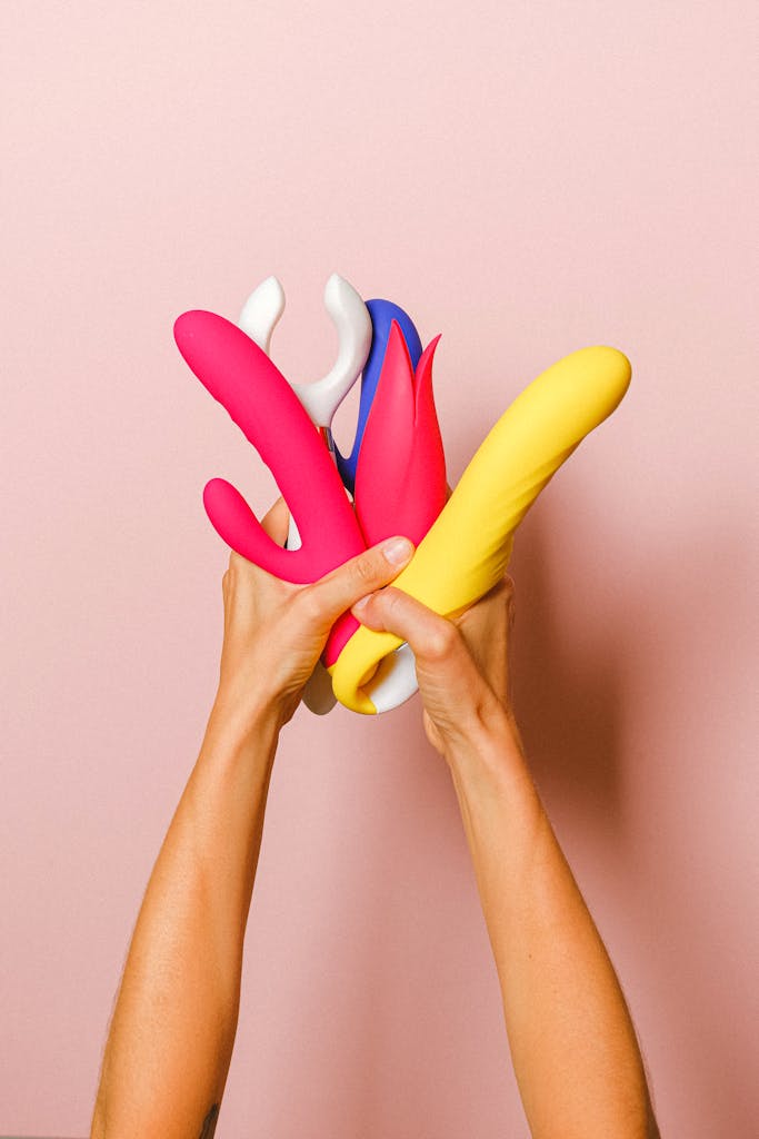 Hands holding vibrant silicone vibrators, showcasing diverse designs against a pink backdrop.