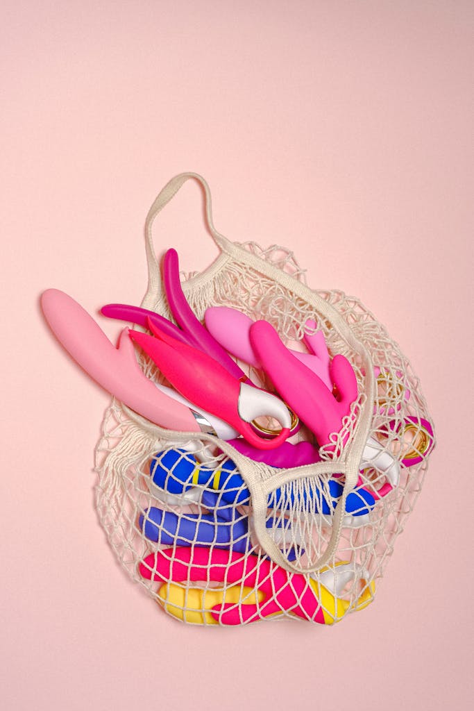 A variety of bright sex toys in a mesh bag on a soft pink background, showcasing modern sensuality.